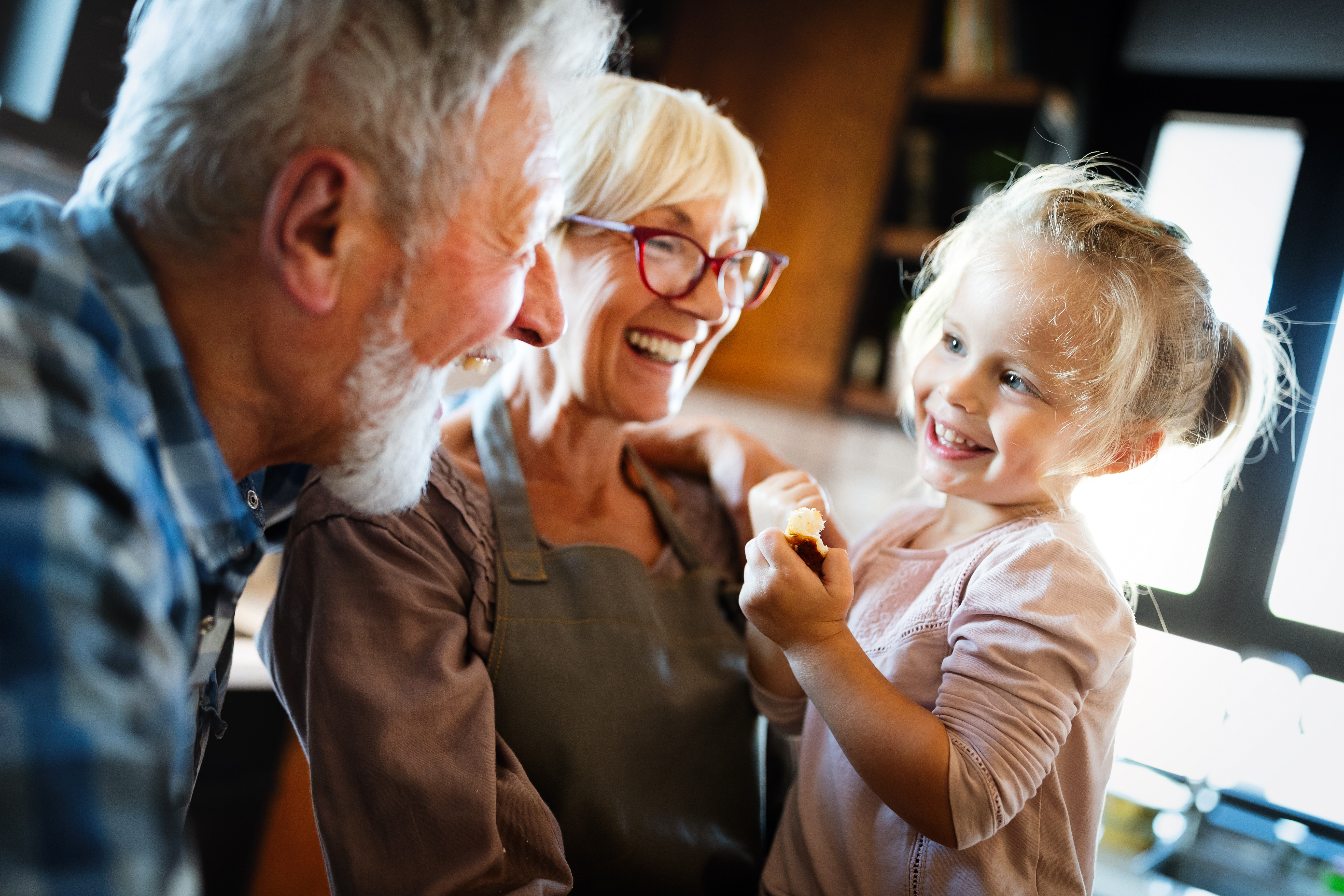 Grandparents laugh with a young granddaughter in a sunlit kitchen