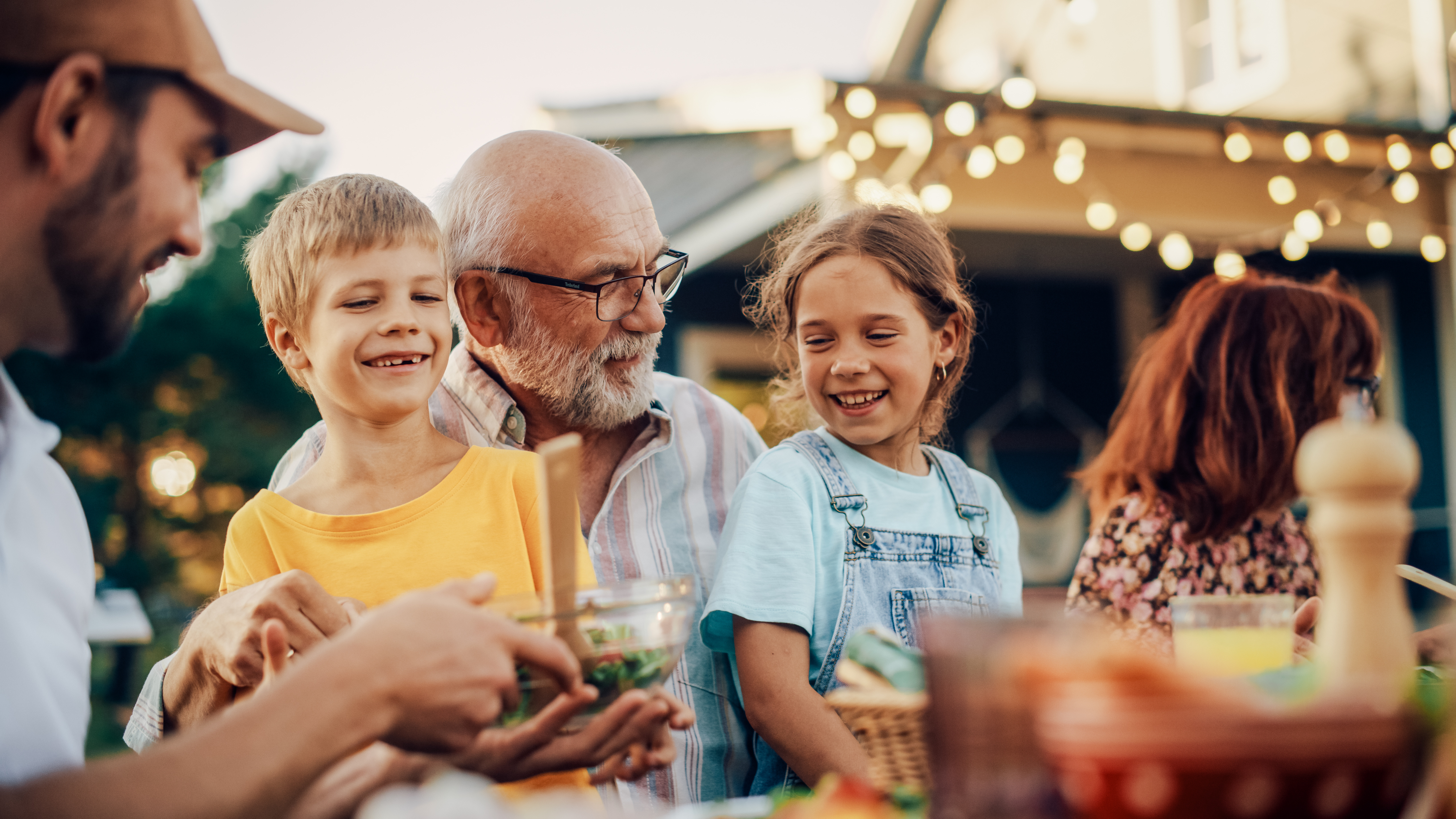 A grandfather and two children smile around an outdoor family table under string lights