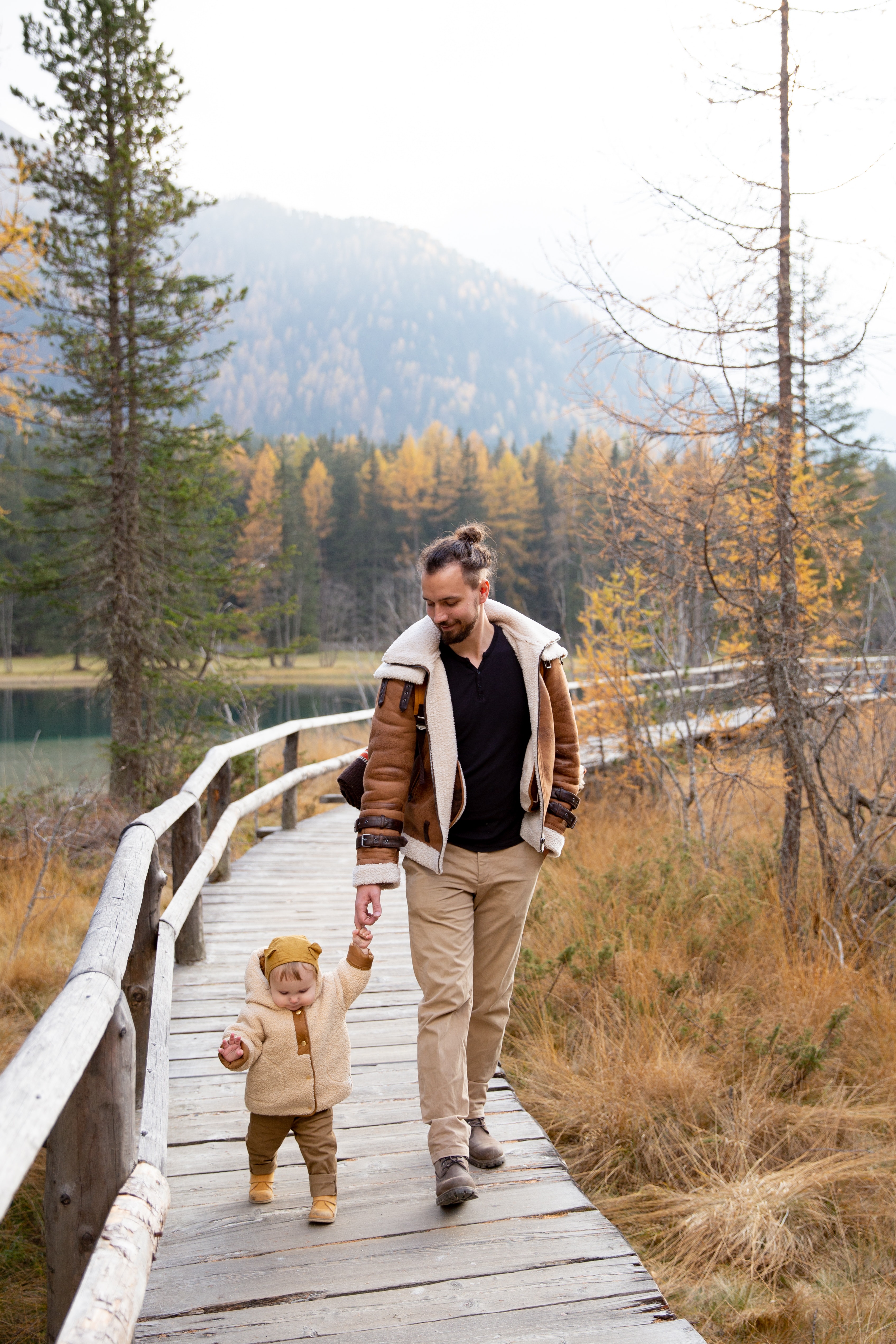 A father and toddler walk a wooden lakeside boardwalk through autumn trees